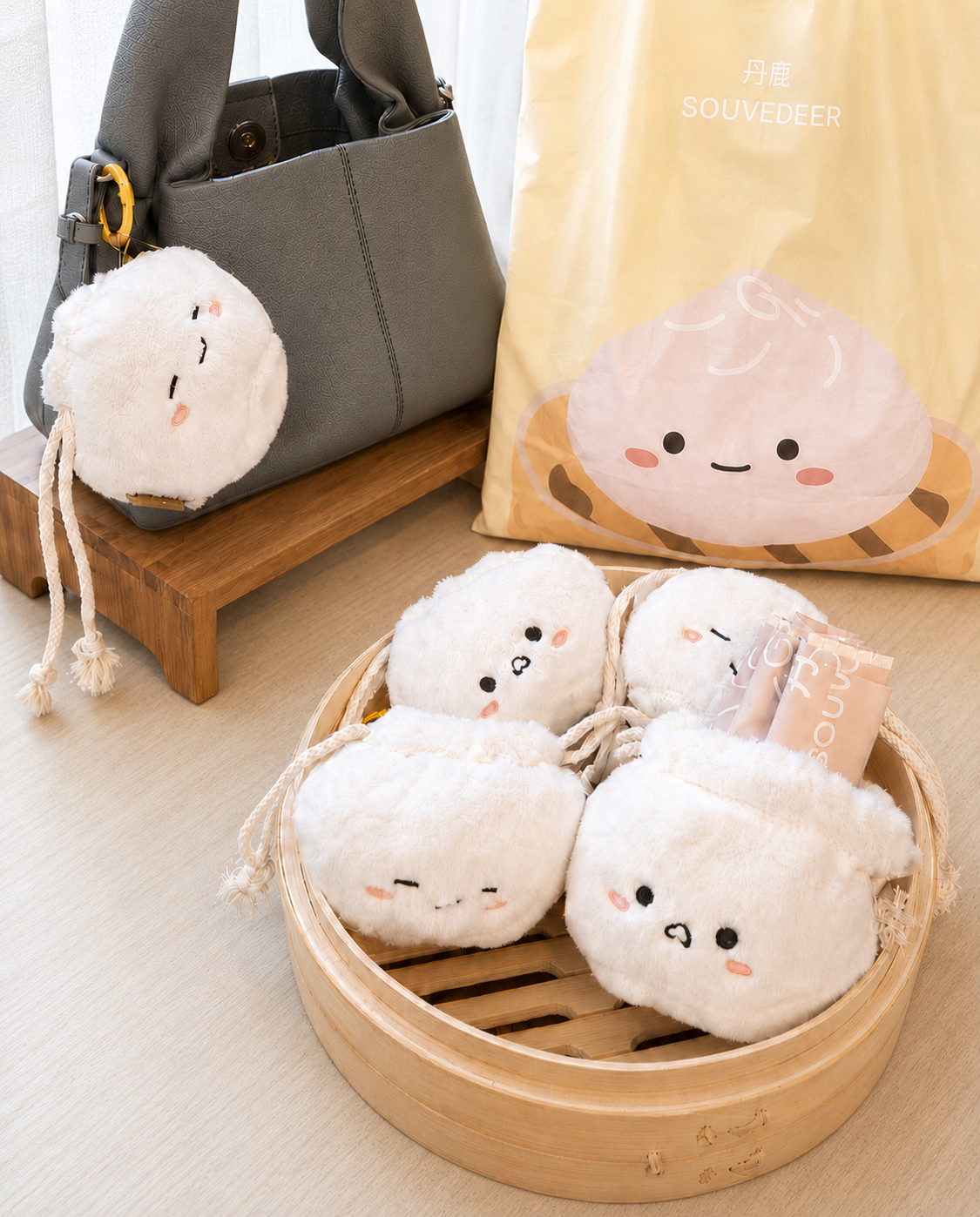 Four plush drawstring pouches shaped like cute white dumplings with embroidered facial features are displayed in a bamboo steamer basket, with a gray handbag and a yellow Souyeer bag in the background.