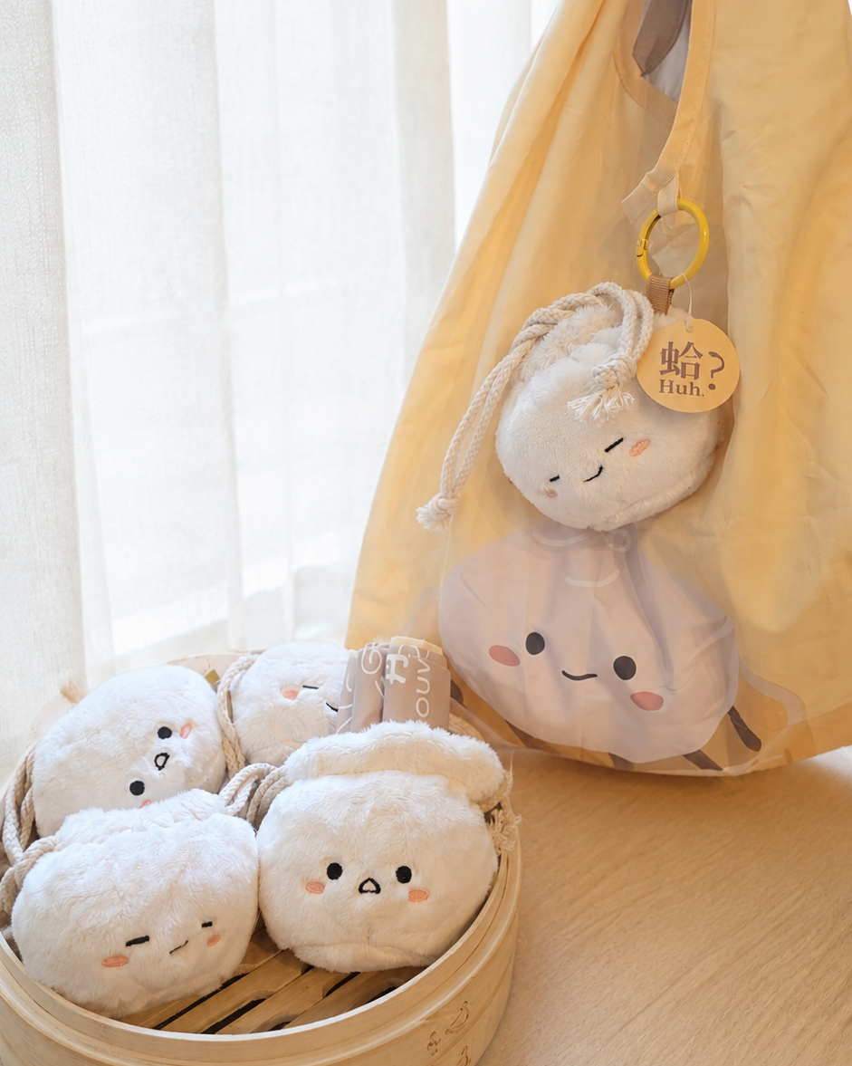 A set of cute, round, plush white characters with embroidered facial features, shown in a bamboo steamer basket and hanging from a yellow tote bag on a light wooden table.
